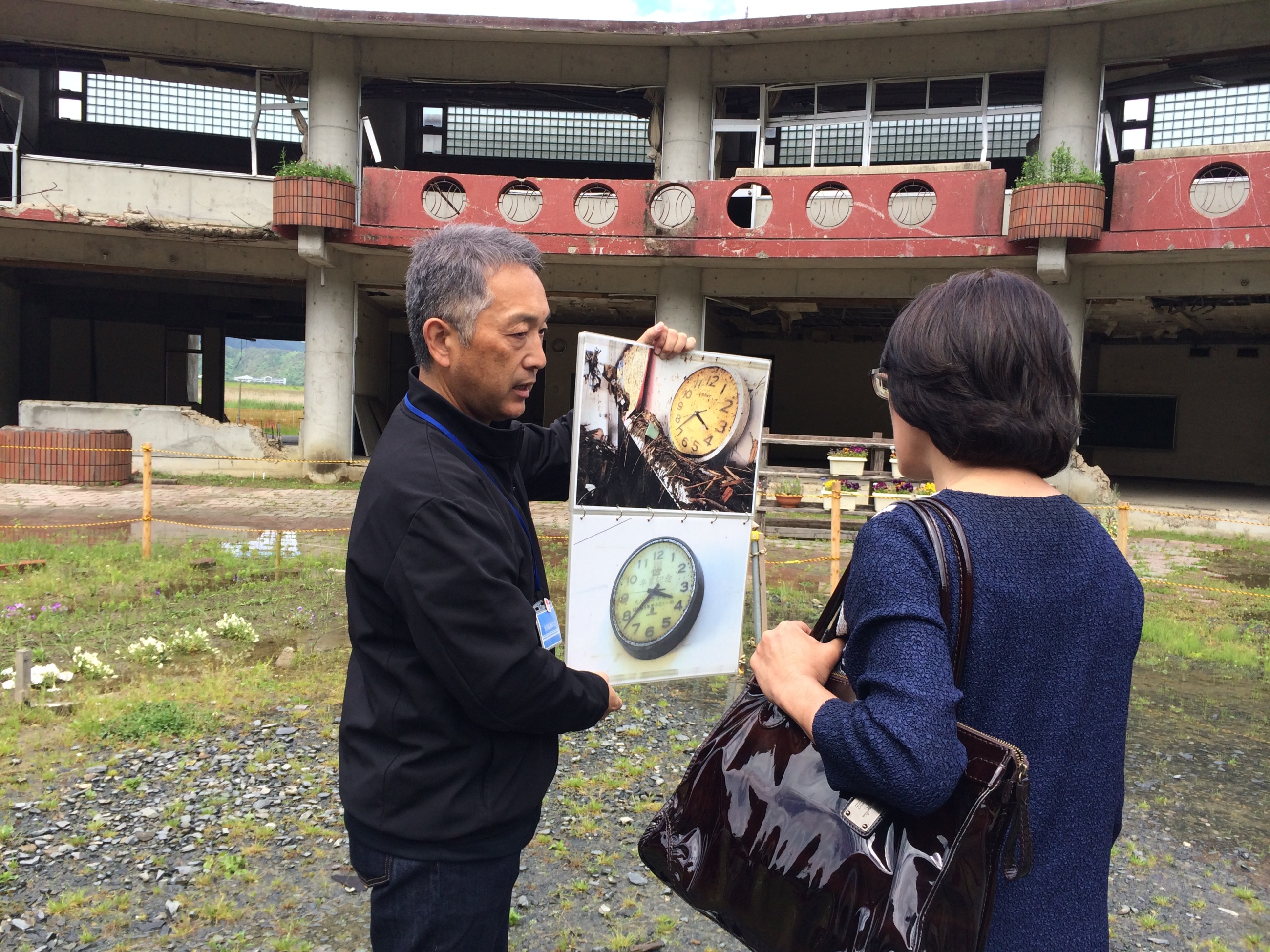 津波で大きな被害を受けた宮城県石巻市立大川小学校の跡地で、水鳥さん（右）が、津波が到達した時間で止まった時計の写真を見ています＝2018年、国連防災機関（UNDRR）提供